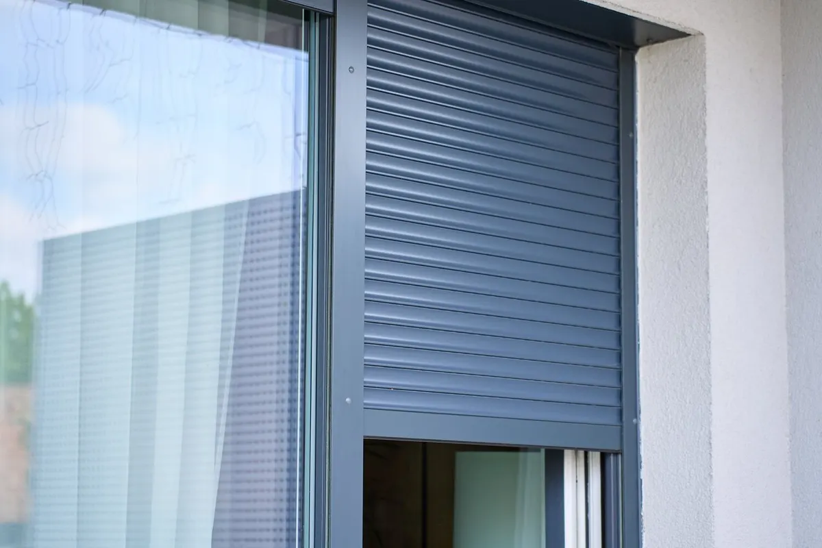 Volet roulant à lames aluminium gris anthracite à moitié descendu devant une fenêtre, dans une façade enduite claire