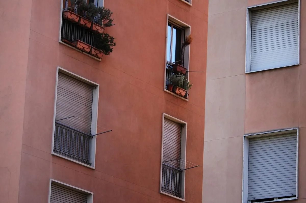 Façade d'immeuble enduite rose terre et ocre, fenêtres équipées de volets roulants blancs à demi baissés, jardinières aux balcons